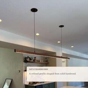 May include: A linear pendant light fixture featuring a long, horizontal bar made of dark brown solid hardwood with softly rounded ends. The fixture is suspended from the ceiling by two thin black cables and includes an integrated LED light strip.