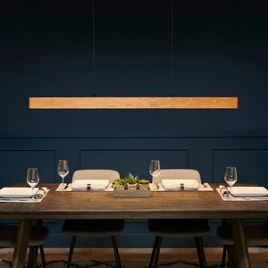 Op de afbeelding: Een rechthoekige hanglamp van hout hangt boven een donkerhouten eettafel met vier stoelen. De tafel is gedekt met placemats, borden en glazen. De kamer heeft donkerblauwe muren en een tapijt met een patroon.