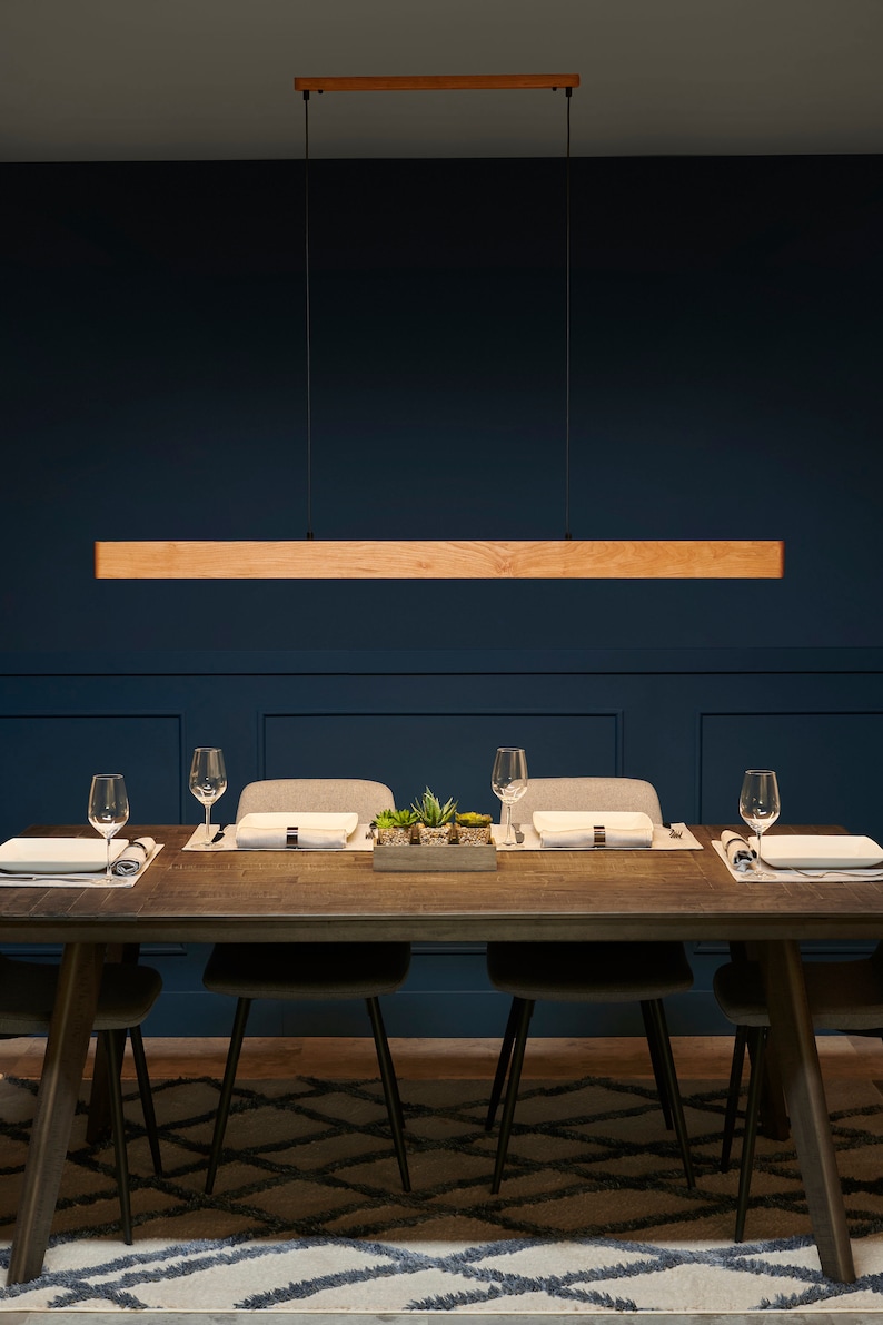 Op de afbeelding: Een rechthoekige hanglamp van hout hangt boven een donkerhouten eettafel met vier stoelen. De tafel is gedekt met placemats, borden en glazen. De kamer heeft donkerblauwe muren en een tapijt met een patroon.
