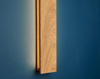 May include: A rectangular wall sconce featuring a vertical light oak wood panel with a prominent natural grain pattern. The fixture emits a warm, soft glow from the left side, casting light against the wall surface behind the wooden structure.