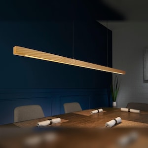 May include: A linear pendant light fixture featuring a long, horizontal wooden beam with a natural grain finish. The fixture is suspended by thin wires and contains an integrated light source that casts a warm glow downward onto the table below.