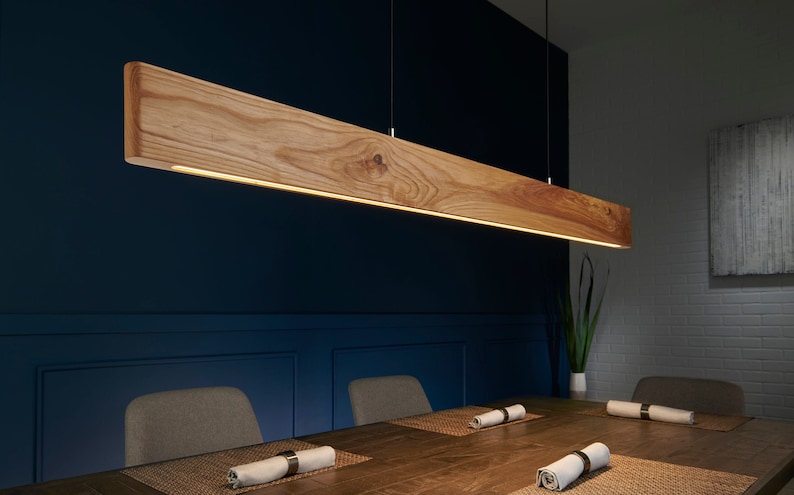May include: A wooden pendant light fixture with a warm, natural wood finish hangs over a dining table. The light fixture is suspended from the ceiling by thin wires. The table is made of wood and has a light brown finish. There are two chairs on either side of the table, and a plant is visible in the background.