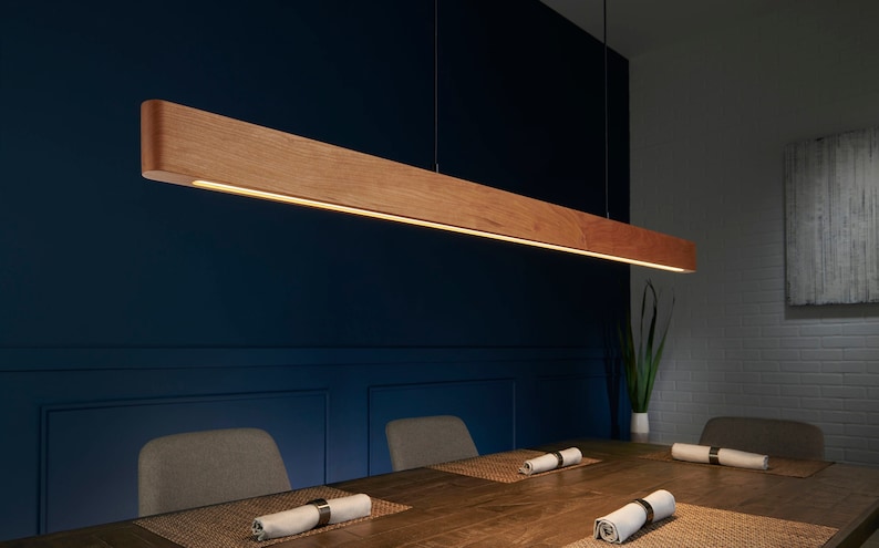 Op de afbeelding: Een rechthoekige houten hanglamp met een warm wit lichtbron hangend boven een houten tafel met twee stoelen. De lamp is aan een dunne draad aan het plafond gehangen.