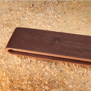 May include: A pair of dark brown wooden tongs with a smooth, curved handle design. The tongs feature a layered construction with a lighter wood tone visible on the inner edges, showcasing a clean, minimalist aesthetic and a polished finish.
