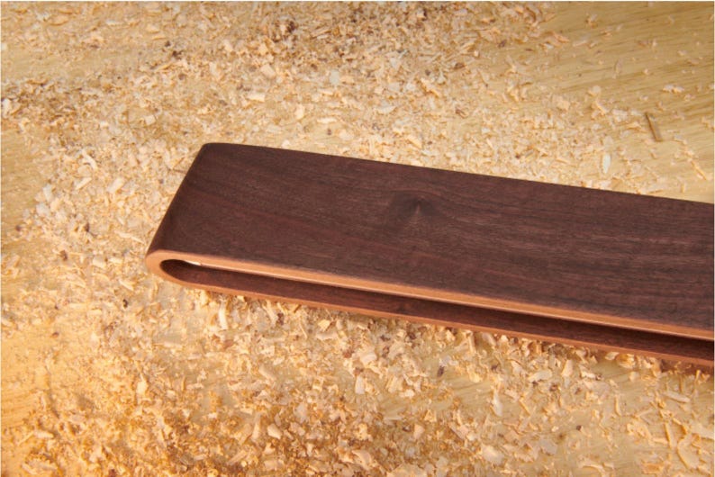 May include: A pair of dark brown wooden tongs with a smooth, curved handle design. The tongs feature a layered construction with a lighter wood tone visible on the inner edges, showcasing a clean, minimalist aesthetic and a polished finish.