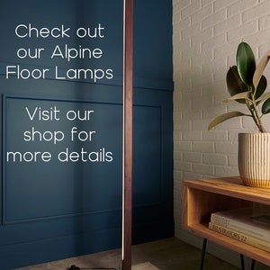 May include: A modern floor lamp with a wooden base and a long, thin light source. The lamp is standing on a rug with a geometric pattern. The lamp is in a room with a white brick wall and a wooden table with books on it.