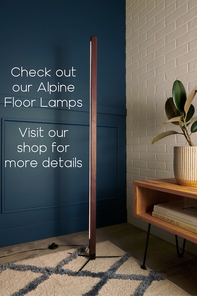 May include: A modern floor lamp with a wooden base and a long, thin light source. The lamp is standing on a rug with a geometric pattern. The lamp is in a room with a white brick wall and a wooden table with books on it.