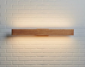 Puede incluir: Un aplique de pared rectangular horizontal hecho de madera marrón natural con un patrón de veta visible. La lámpara emite un brillo cálido y suave desde los bordes superior e inferior, creando un efecto de luz difusa en la pared.