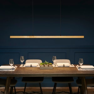 May include: A wooden rectangular pendant light fixture hangs over a dark wood dining table with four chairs. The table is set with placemats, plates, and glasses. The room has dark blue walls and a patterned rug.
