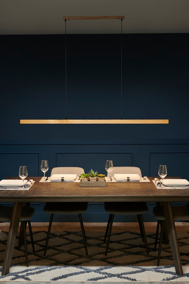 May include: A wooden rectangular pendant light fixture hangs over a dark wood dining table with four chairs. The table is set with placemats, plates, and glasses. The room has dark blue walls and a patterned rug.
