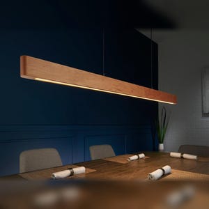 May include: A long, linear pendant light fixture crafted from natural wood with a smooth finish. The rectangular beam features an integrated LED light strip running along the bottom edge, suspended by thin wires from above the dining table.