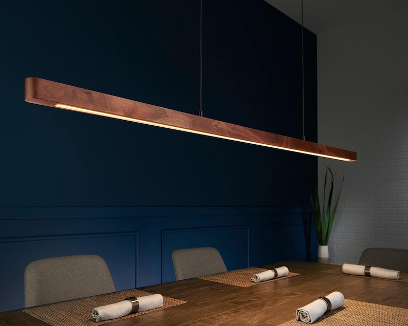 May include: A long, linear pendant light fixture crafted from dark brown wood with a smooth finish. The fixture is suspended by thin wires and features an integrated LED light strip running along the bottom edge, emitting a warm, downward glow.