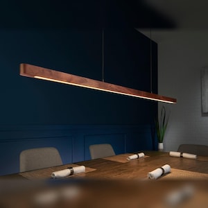 May include: A long, linear pendant light fixture made of dark brown wood with a slim, integrated LED light strip running along the bottom edge. The fixture is suspended by thin wires above a wooden dining table in a dimly lit room.