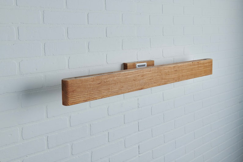 May include: A wooden wall-mounted shelf with a natural wood finish. The shelf is mounted on a white brick wall.