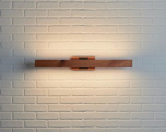 Puede incluir: Un aplique de pared horizontal que presenta una pieza larga y rectangular de madera marrón natural con veta visible. La lámpara incluye un soporte central de metal oscuro y emite un brillo suave y cálido desde detrás de la madera.