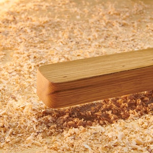 May include: A close-up of a wooden plank resting on a bed of wood shavings. The plank is light brown and has a smooth, polished surface.