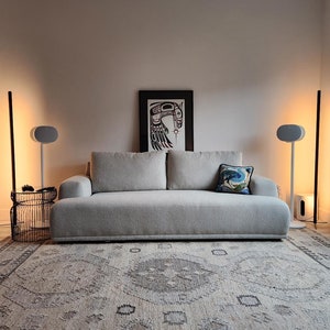 Peut inclure: Un canap&eacute; d'angle gris avec un coussin d&eacute;coratif &agrave; motifs est plac&eacute; sur un tapis beige dans un salon. Deux enceintes blanches sont sur des supports de chaque c&ocirc;t&eacute; du canap&eacute;. Un ventilateur de plafond noir est au-dessus du canap&eacute;. Une estampe encadr&eacute;e d'un motif noir et rouge est accroch&eacute;e au mur derri&egrave;re le canap&eacute;.