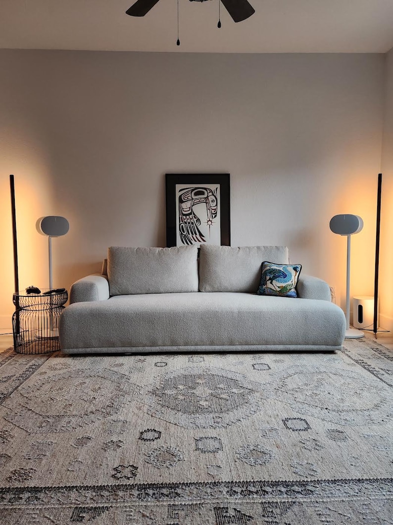 Peut inclure: Un canap&eacute; d'angle gris avec un coussin d&eacute;coratif &agrave; motifs est plac&eacute; sur un tapis beige dans un salon. Deux enceintes blanches sont sur des supports de chaque c&ocirc;t&eacute; du canap&eacute;. Un ventilateur de plafond noir est au-dessus du canap&eacute;. Une estampe encadr&eacute;e d'un motif noir et rouge est accroch&eacute;e au mur derri&egrave;re le canap&eacute;.