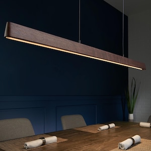 May include: A linear pendant light fixture featuring a long, dark brown wooden beam with a smooth finish. The fixture is suspended by thin wires from the ceiling and emits a warm, continuous strip of light downward from its underside.