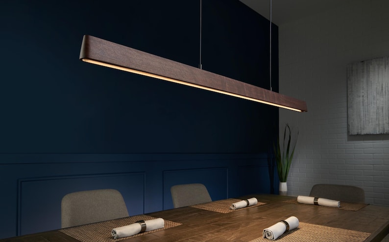 May include: A linear pendant light fixture featuring a long, dark brown wooden beam with a smooth finish. The fixture is suspended by thin wires from the ceiling and emits a warm, continuous strip of light downward from its underside.