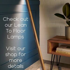 May include: A wooden floor lamp with a long, slender design. The lamp is leaning against a wall and casting a warm light. The lamp is on a rug with a geometric pattern. The text "Check out our Lean To Floor Lamps Visit our shop for more details" is visible in the image.