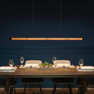 Pu&ograve; includere: Un apparecchio di illuminazione lungo e rettangolare in legno &egrave; appeso sopra un tavolo da pranzo con quattro sedie. Il tavolo &egrave; in legno scuro e ha un tappeto bianco sotto. Le pareti sono di colore blu scuro.