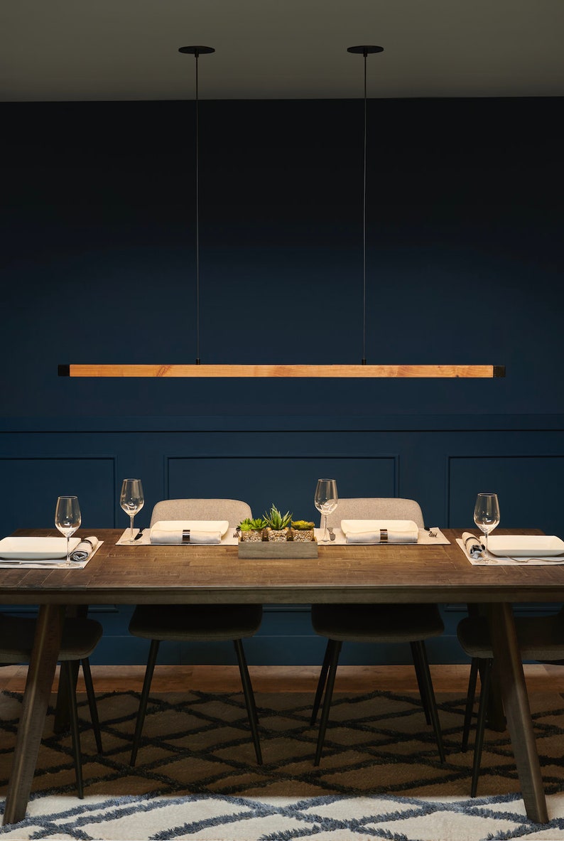 Pu&ograve; includere: Un apparecchio di illuminazione lungo e rettangolare in legno &egrave; appeso sopra un tavolo da pranzo con quattro sedie. Il tavolo &egrave; in legno scuro e ha un tappeto bianco sotto. Le pareti sono di colore blu scuro.