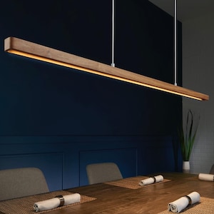 May include: A linear pendant light fixture featuring a long, rectangular wooden housing with a warm, integrated LED light strip running along the bottom edge, suspended from the ceiling by two thin white cables over a dining table.