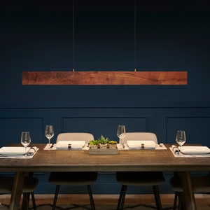 May include: A wooden rectangular pendant light fixture hangs over a dining table with four chairs. The table is set with placemats, plates, and glasses. The room has dark blue walls and a patterned rug.