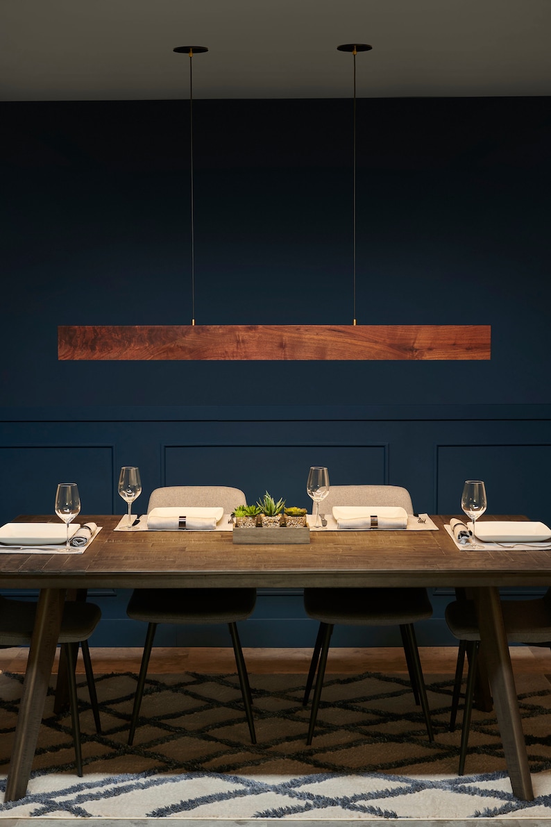 May include: A wooden rectangular pendant light fixture hangs over a dining table with four chairs. The table is set with placemats, plates, and glasses. The room has dark blue walls and a patterned rug.
