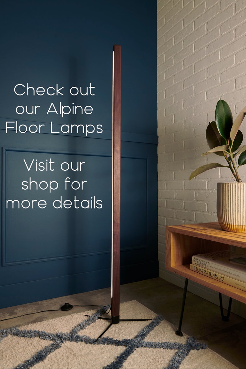 May include: A modern floor lamp with a wooden base and a long, thin light source. The lamp is standing on a rug with a geometric pattern. The lamp is in a room with a white brick wall and a wooden table with books on it.