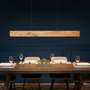 May include: A wooden rectangular pendant light fixture hangs over a dining table with four place settings. The table has a dark wood finish and is set with white plates, silverware, and glasses. The table is on a patterned rug in a room with dark blue walls.