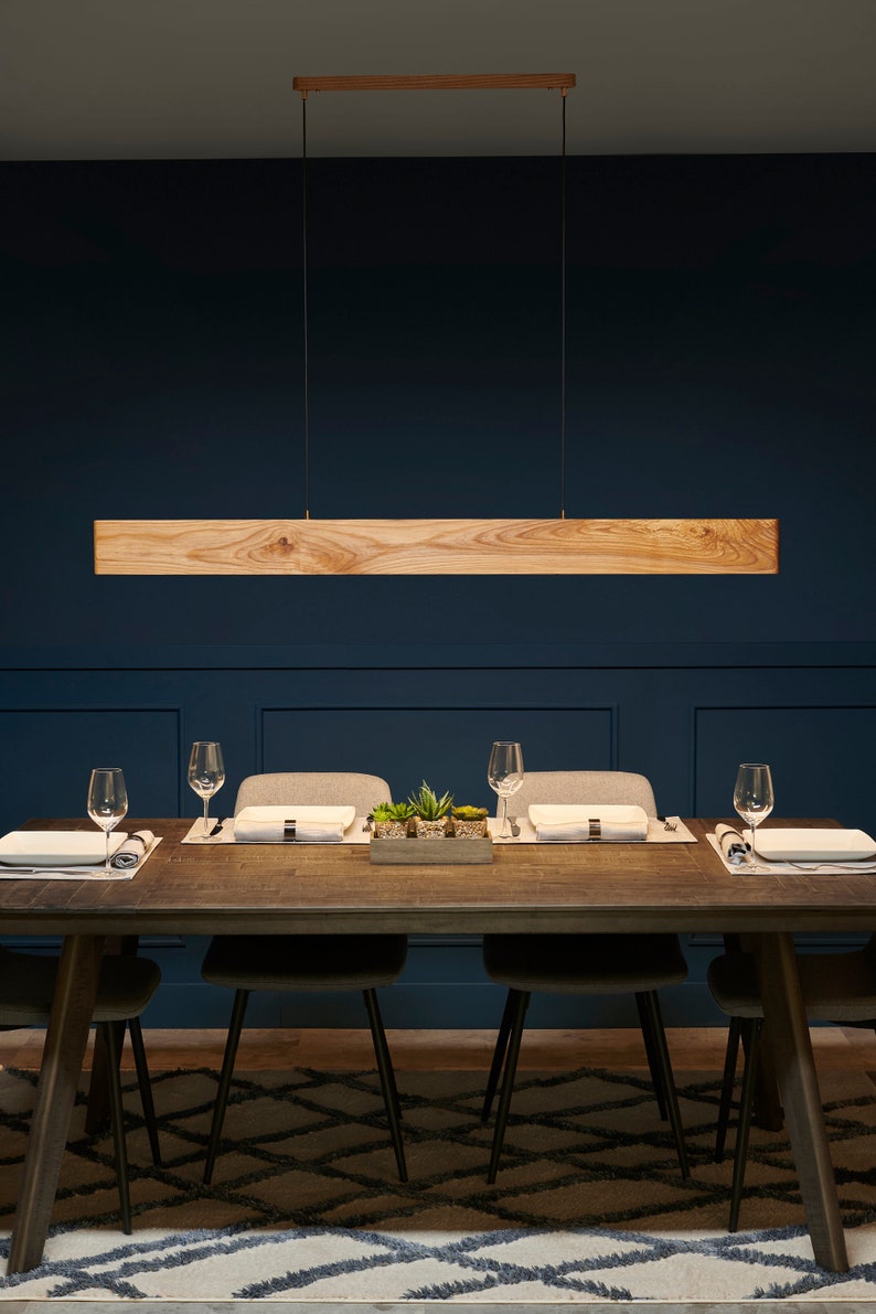 May include: A wooden rectangular pendant light fixture hangs over a dining table with four place settings. The table has a dark wood finish and is set with white plates, silverware, and glasses. The table is on a patterned rug in a room with dark blue walls.
