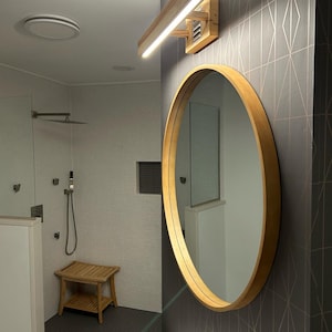 May include: A modern bathroom with a round wooden framed mirror, a white vanity sink, and a wooden light fixture with a white light strip. The bathroom has a shower with a glass door and a tiled floor.