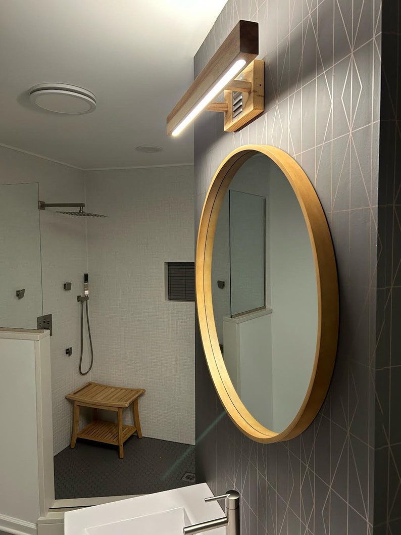 May include: A modern bathroom with a round wooden framed mirror, a white vanity sink, and a wooden light fixture with a white light strip. The bathroom has a shower with a glass door and a tiled floor.