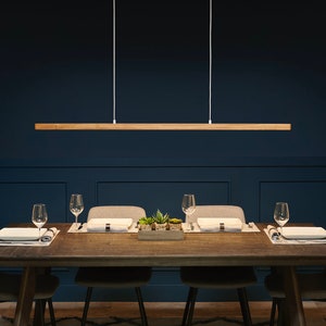 May include: A wooden rectangular pendant light fixture hangs over a dark blue dining room table with four chairs. The table is set with placemats, plates, and glasses.