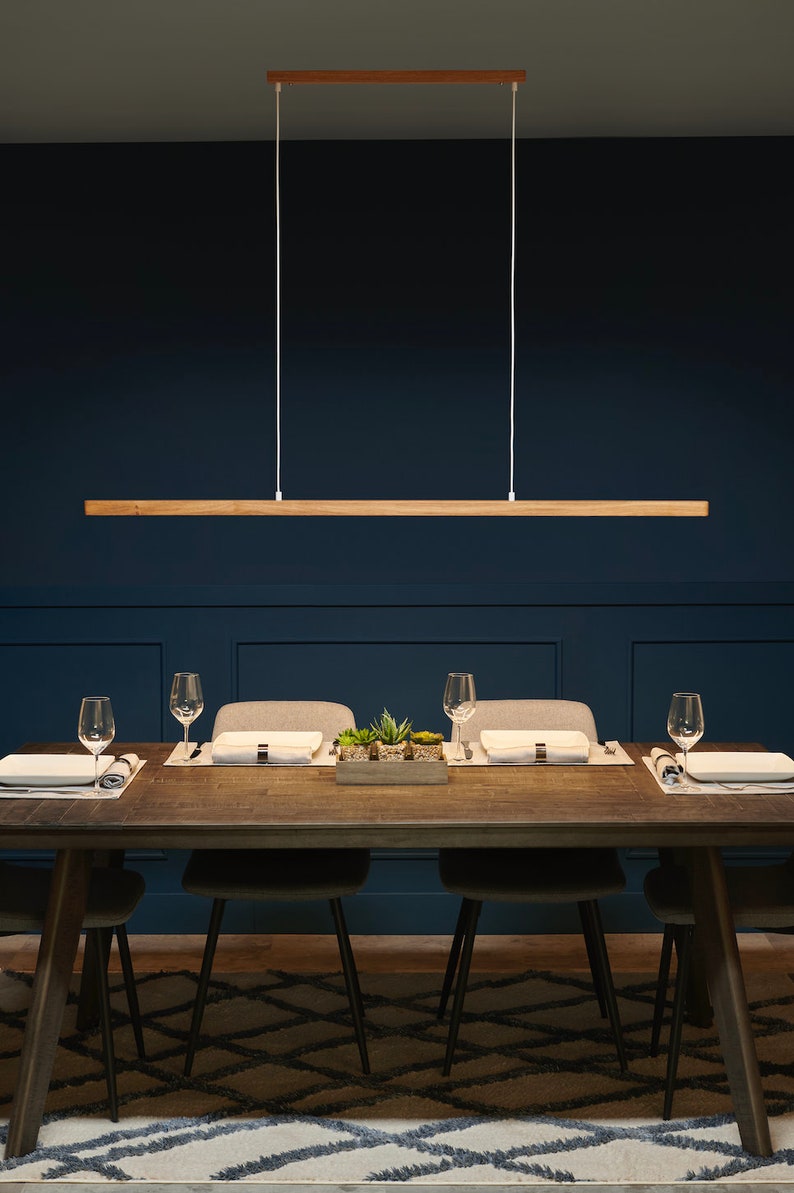 May include: A wooden rectangular pendant light fixture hangs over a dark blue dining room table with four chairs. The table is set with placemats, plates, and glasses.