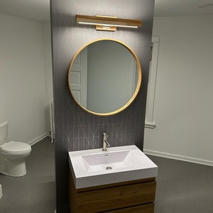 May include: A white bathroom vanity with two drawers and a white porcelain sink. The vanity is mounted on a wall with a gray geometric patterned wallpaper. A round mirror with a gold frame is above the sink. A silver faucet is attached to the sink.