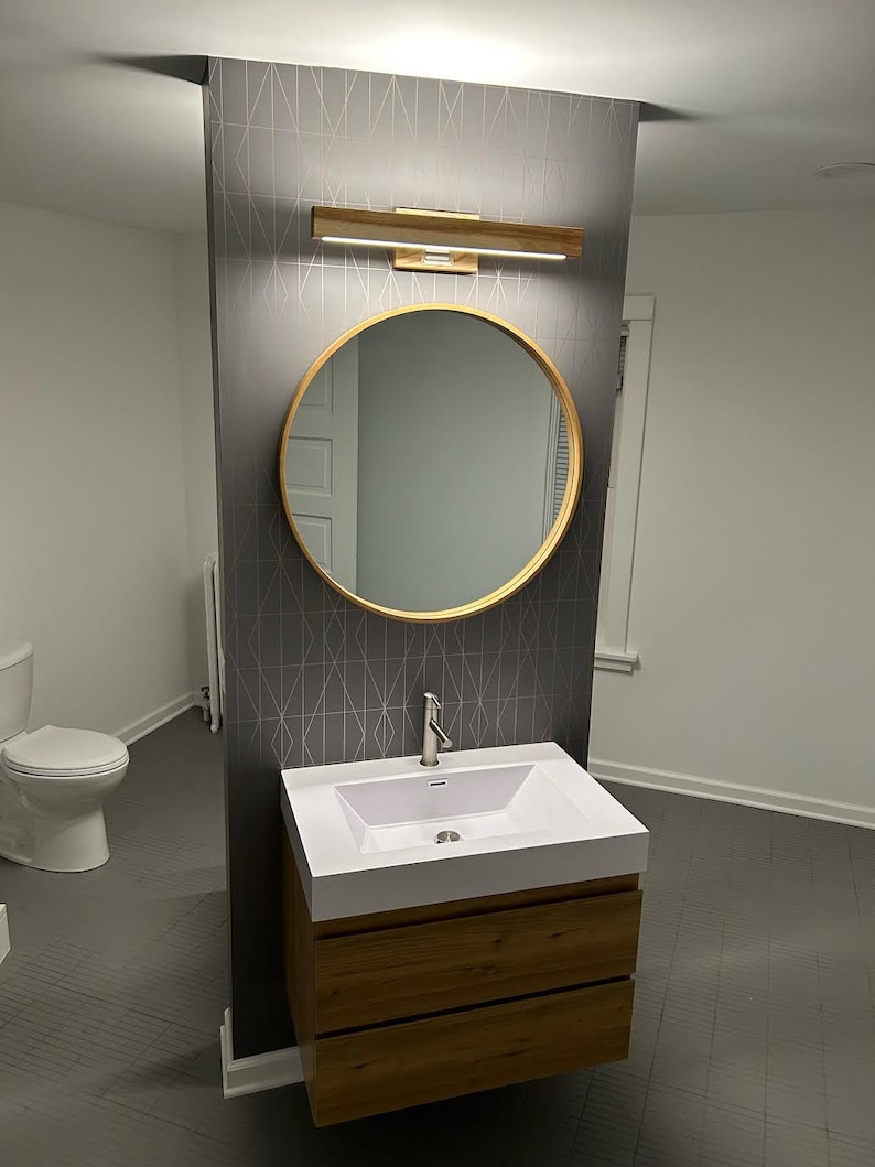 May include: A white bathroom vanity with two drawers and a white porcelain sink. The vanity is mounted on a wall with a gray geometric patterned wallpaper. A round mirror with a gold frame is above the sink. A silver faucet is attached to the sink.
