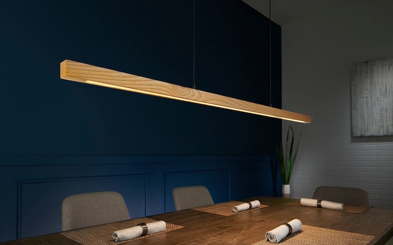 May include: A wooden pendant light fixture hangs over a dining table with two chairs. The light fixture is a long, rectangular piece of wood with a light strip running along the bottom. The table is made of wood and has a brown finish. The chairs are upholstered in a light gray fabric.