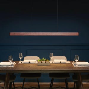 May include: A linear pendant light fixture featuring a long, horizontal wooden beam suspended by two thin black cables from a matching rectangular wooden ceiling mount, providing a clean and straightforward lighting design for a dining area.