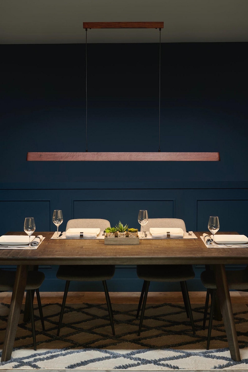 May include: A linear pendant light fixture featuring a long, horizontal wooden beam suspended by two thin black cables from a matching rectangular wooden ceiling mount, providing a clean and straightforward lighting design for a dining area.
