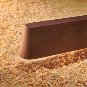 May include: A smooth, dark brown wooden board with a rounded, tapered end. The piece of wood has a matte finish and a straight edge, positioned horizontally above a surface covered in light-colored wood shavings and sawdust from woodworking.