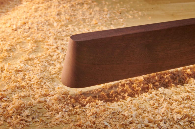 May include: A smooth, dark brown wooden board with a rounded, tapered end. The piece of wood has a matte finish and a straight edge, positioned horizontally above a surface covered in light-colored wood shavings and sawdust from woodworking.