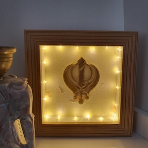 May include: A framed illuminated artwork featuring a gold Khanda, a symbol of Sikhism, on a white background with a string of white lights around the border.