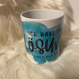 Könnte beinhalten: Weißer Keramikbecher mit blauem Aquarelldesign und schwarzer Schrift. Der Text auf dem Becher lautet "ICH HABE EINE LÖSUNG, ABER DIE PASST NICHT ZUM". Der Becher ist zylindrisch und steht auf einer flauschigen weißen Oberfläche.