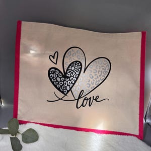 Könnte beinhalten: Eine beigefarbene Tragetasche aus Segeltuch mit leuchtend pinkfarbenen Seitenrändern. Die Vorderseite zeigt eine Grafik aus zwei überlappenden Herzen mit schwarz-silbernem Leopardenmuster, einem kleinen schwarzen Herz und dem Wort love in schwarzer Schreibschrift.