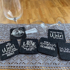 Könnte beinhalten: Sechs schwarze Filzuntersetzer mit weißem Text. Die Untersetzer zeigen Sprüche zum Thema Wein wie "Der beste Wein ist der, den man mit Freunden trinkt" und "Meine Laune ist im Keller, hoffe sie bringt Wein mit."