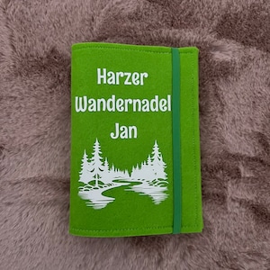 Könnte beinhalten: Grünes Filz-Notizbuch mit einem weißen Design aus Bäumen und einem Fluss. Der Text "Harzer Wandernadel Jan" ist auf der Vorderseite aufgedruckt.