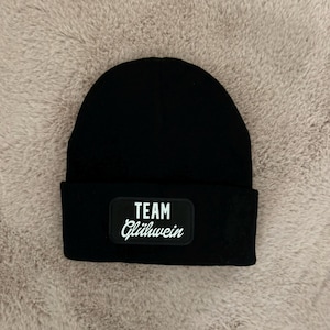 Könnte beinhalten: Eine schwarze Beanie-Mütze mit einem rechteckigen Aufnäher mit dem weißen Schriftzug "TEAM Glühwein". Die Mütze ist aus weichem Strickmaterial gefertigt und für kaltes Wetter geeignet. Die Beanie ist ein Winteraccessoire.